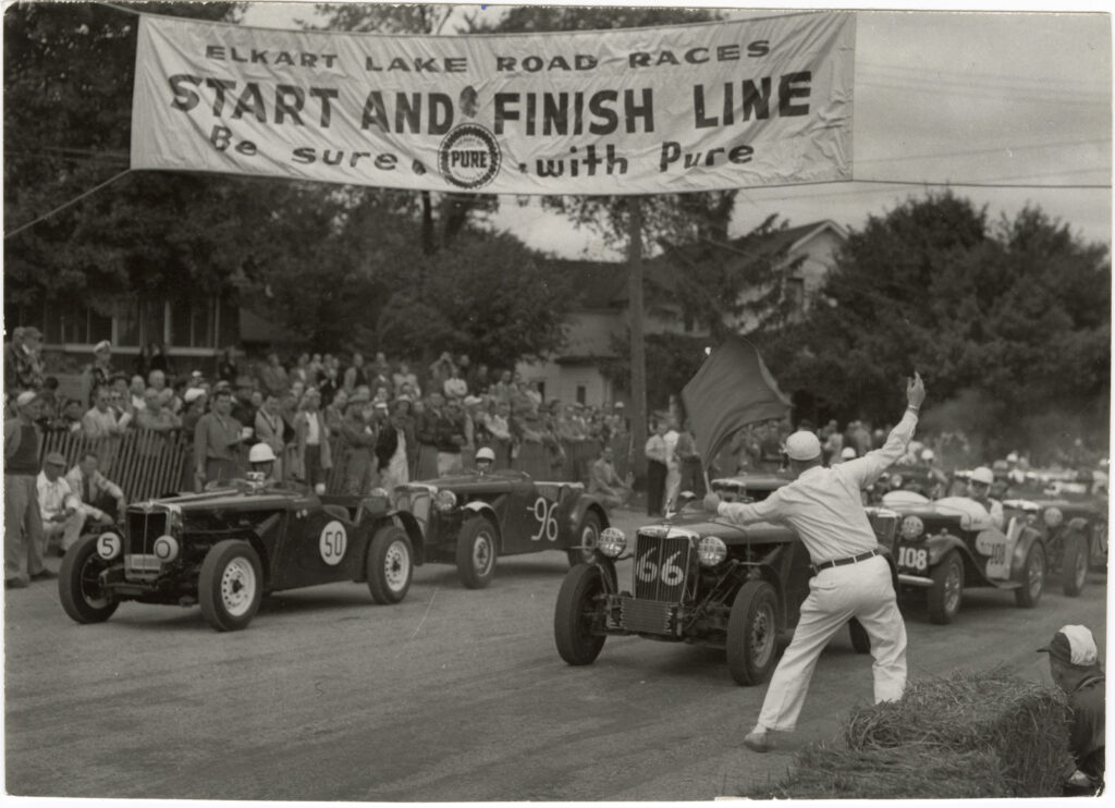 A black and white photo of a man waving a start flag to a car race. A banner above reads "Elkart Lake Road Races. Start and Finish Line. Be sure. With Pure.