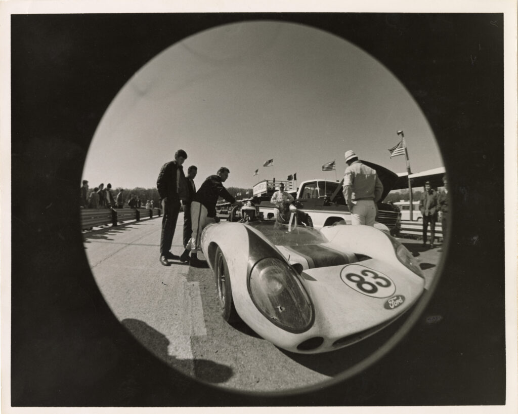 Black and white photo of numerous men looking at a racing car