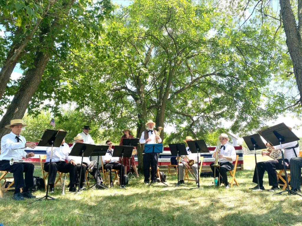 Band playing outdoors in a semi-circle with music stands, all wearing white shirts and hats