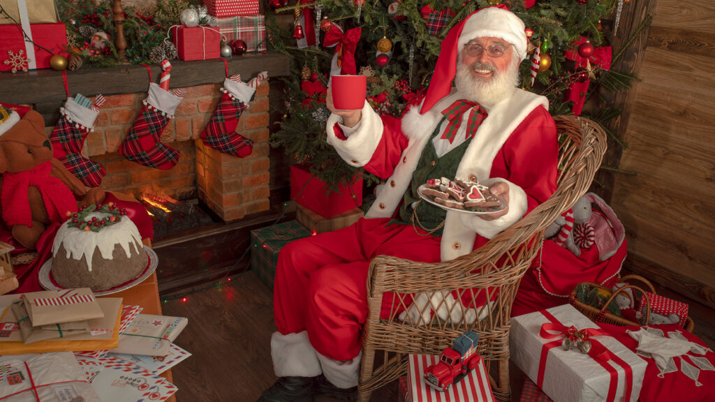 A white man dresses as Santa, holding a red mug and a plate of red and white decorated cookies. He is surrounded by wrapped presents and next to a decorated Christmas tree. 