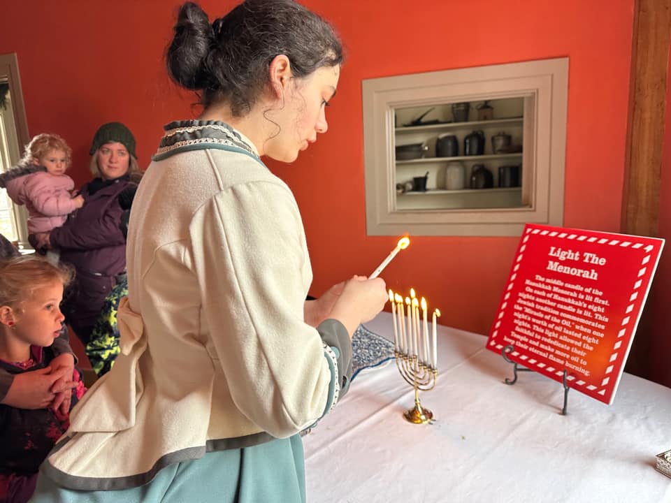A woman with dark colored hair, wearing period time clothing, lights the menorah candles with a group of people watching.