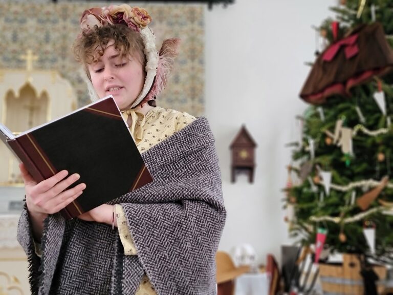 A woman with fair complexion, wearing time period clothing, reading and singing from a dark colored book