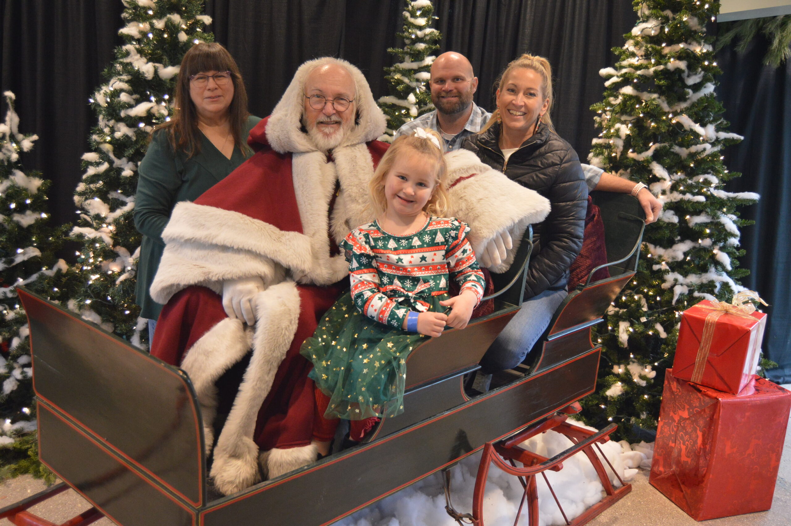 A family photo of three adults and one younger child sitting on a dark colored sled with an older man dressed as Santa.