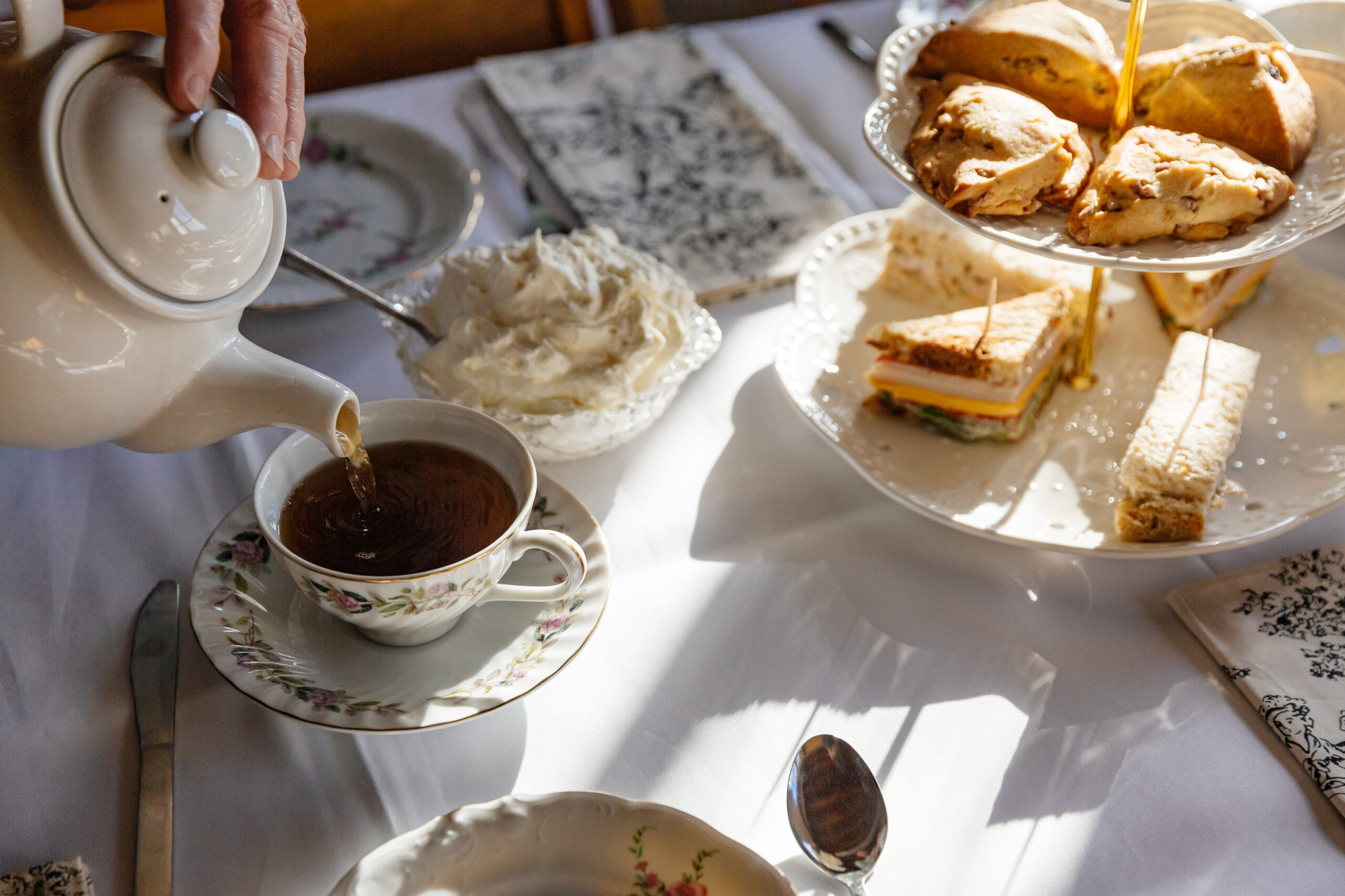 A spread of baked goods and sandwiches on a white tablecloth with someone pouring a cup of tea.